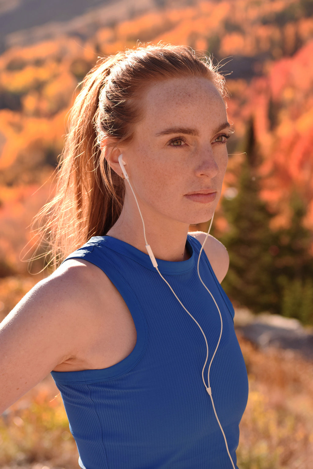 Woman in a blue sleeveless shirt with earphones, standing in a scenic outdoor setting with autumn foliage.
