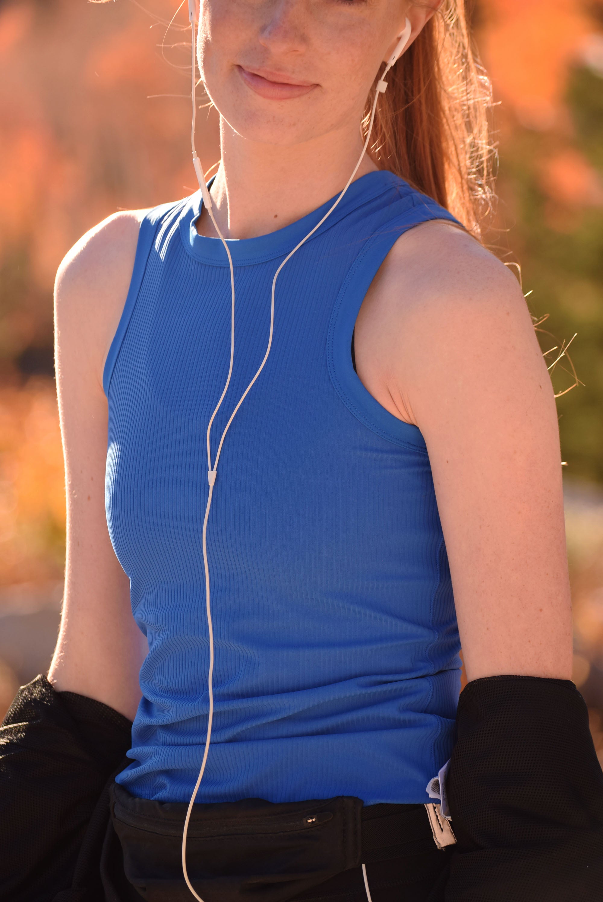 Person wearing a blue sleeveless shirt with earphones, standing outdoors with a blurred background