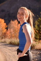 Woman in a blue tank top standing outdoors with autumn foliage in the background
