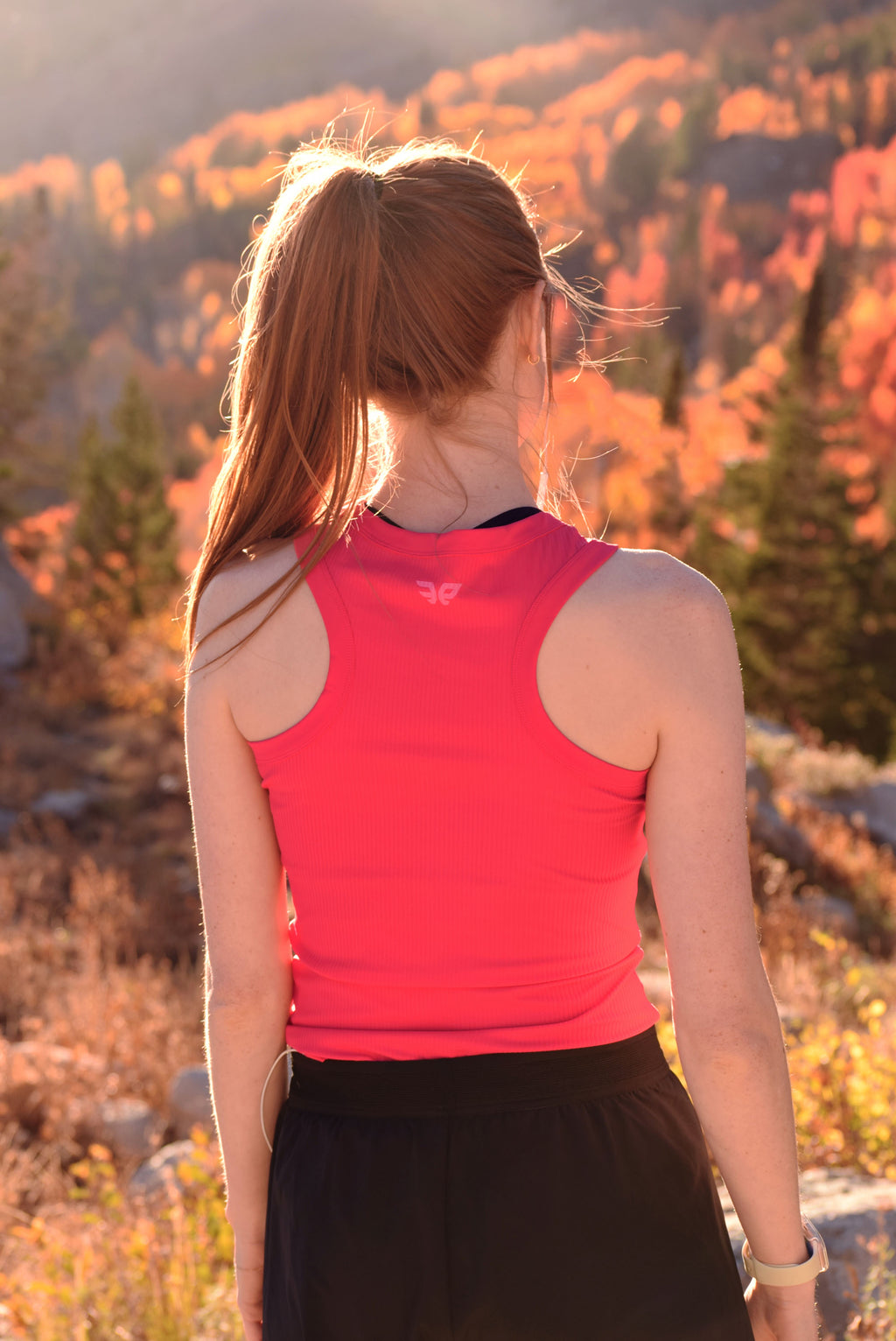 Person wearing a red tank top and black shorts standing in a scenic outdoor setting with trees and mountains.