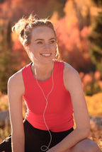 Woman in a red tank top with earphones, sitting outdoors with a blurred natural background