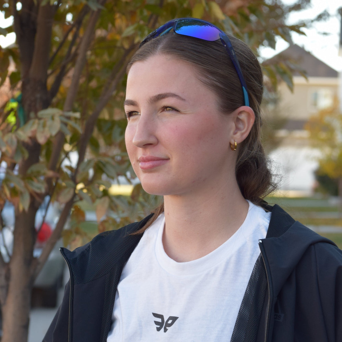 A woman outdoors wearing a white t-shirt with a black logo, a black zip-up hoodie, and blue-tinted sunglasses on her head, with a tree in the background.